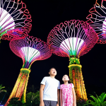 A family (parents and two children) stands in awe, looking up at the illuminated Supertrees during the "Garden Rhapsody" light show at Gardens by the Bay in Singapore. The scene is magical and futuristic, with vibrant colors, lush tropical plants, and a sense of wonder. The children's faces reflect pure amazement.